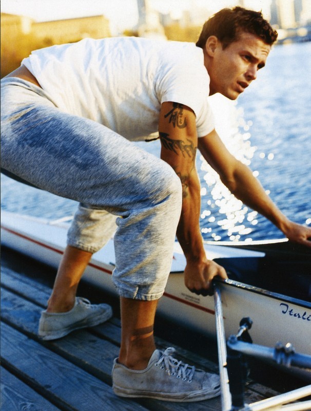 Athletic man wearing a white T-shirt and sweatpants on a wooden dock, holding a rowing shell, with a sunlit waterfront background.