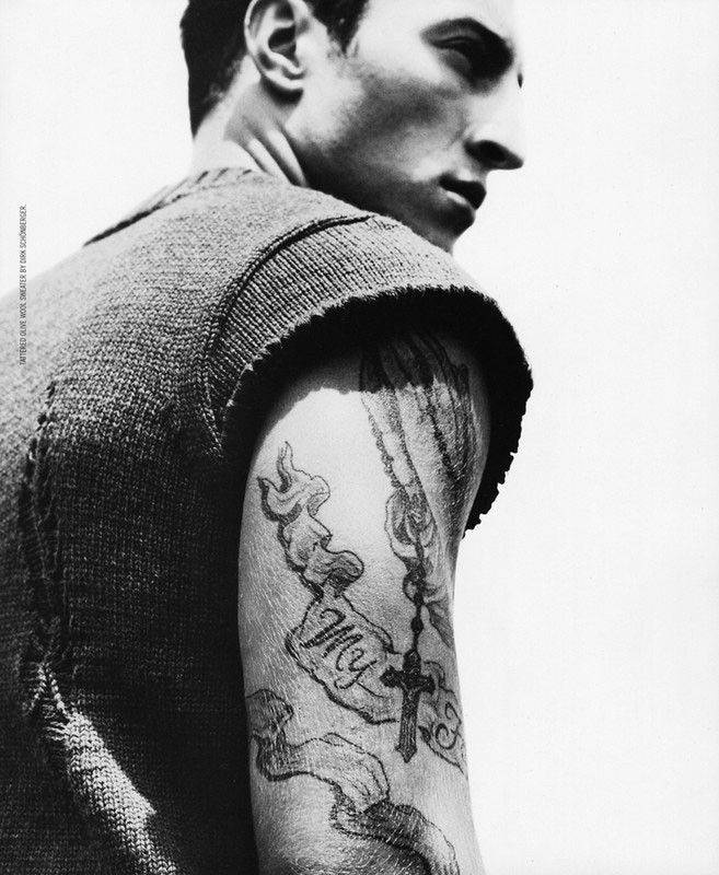 Monochrome portrait of a man with a tattoo, wearing a sweater vest, against a plain background, showcasing shoulder and upper arm.