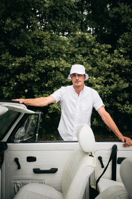 Man in a white bucket hat and shirt stands next to a white convertible car with a green foliage background on an overcast day.