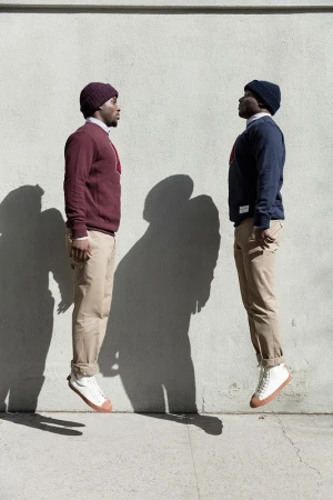 Two African American men in sweaters, hats, and khaki pants appear to float mid-air outdoors against a light grey concrete wall backdrop.