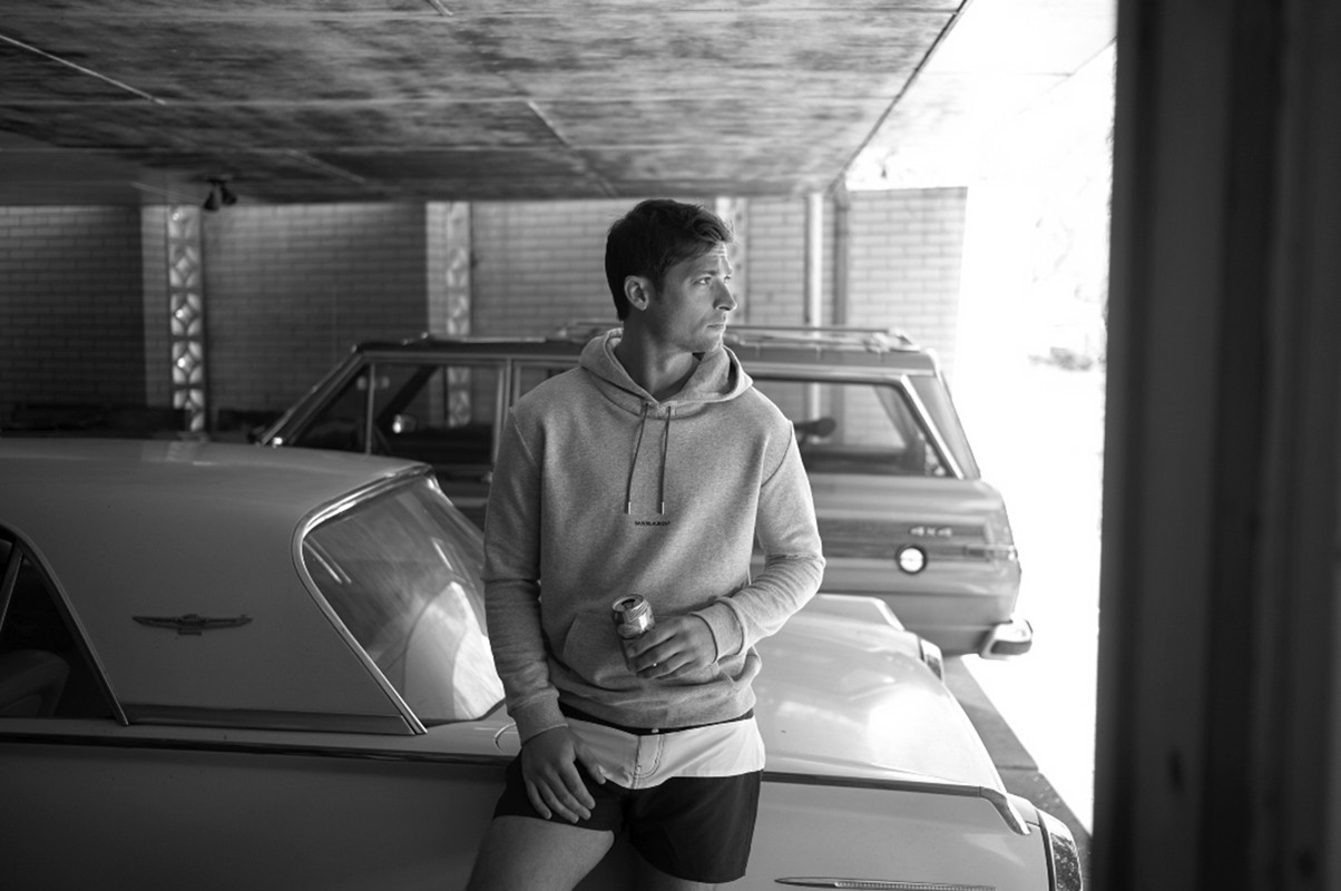 Monochrome shot of a man in a hoodie leaning on a car, holding a can, with another classic car in the garage setting.
