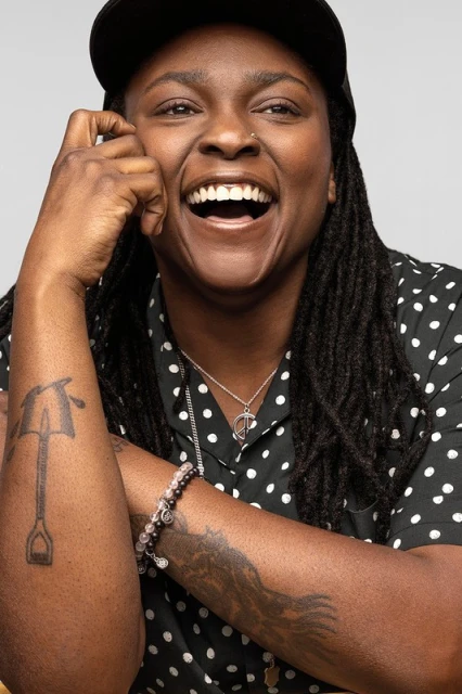 Smiling African-American woman in a black and white polka dot shirt and black baseball cap poses in a studio with tattoos on her arms.