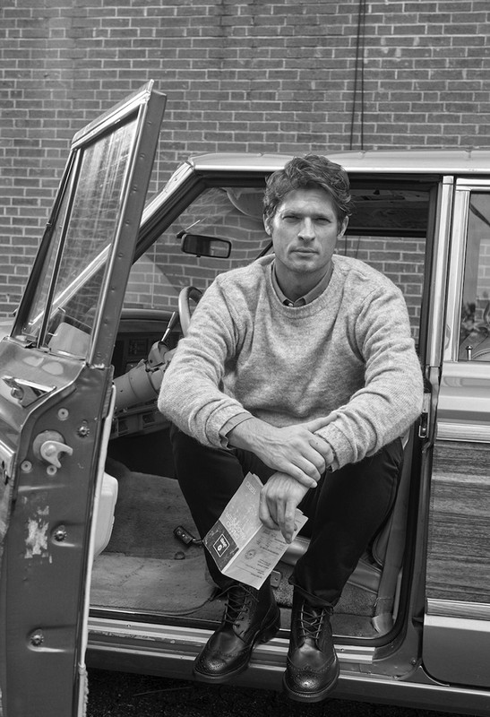 Man sitting in a classic wood-trimmed station wagon, holding papers, wearing a grey sweater and black boots with a brick background.