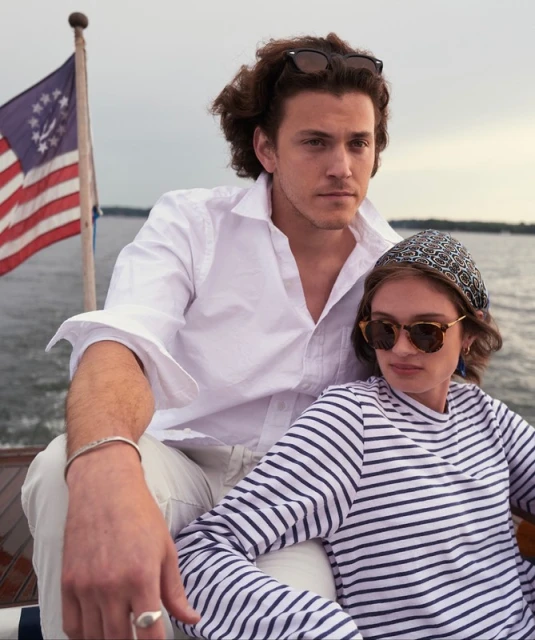 A young couple is on a boat, wearing sunglasses, with the man in a white shirt and the woman in a striped shirt, and a US flag.