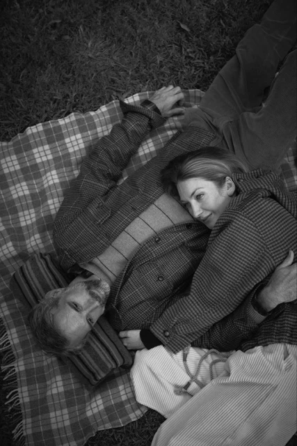 Couple lying on a plaid blanket in a grassy field. Woman embraces man, top-down shot showing checkered jackets, striped shirts, and casual pants.