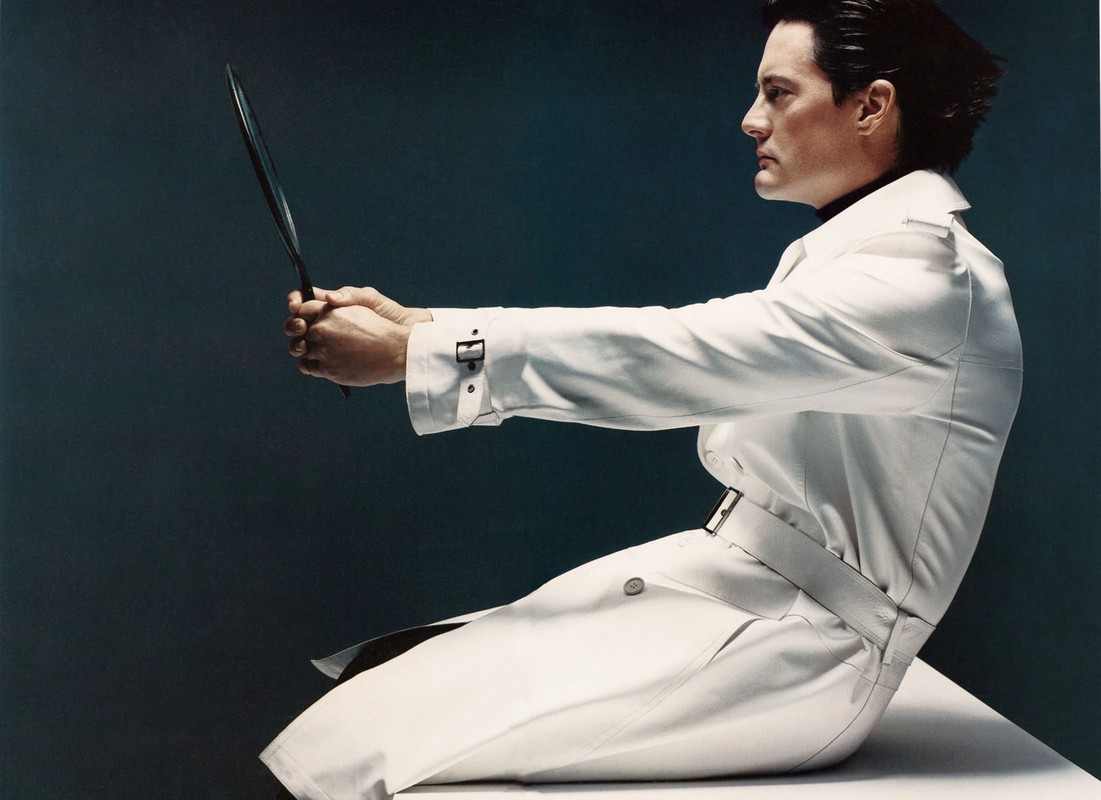 Profile view of an adult male in a white trench coat holding a long, thin object against a dark teal background in a studio shot.