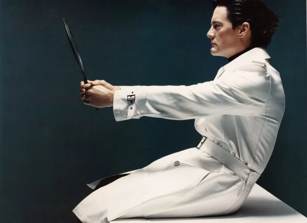 Profile view of an adult male in a white trench coat holding a long, thin object against a dark teal background in a studio shot.