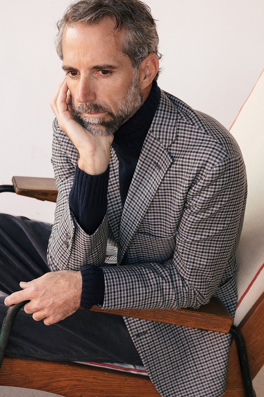 Portrait of a middle-aged man with salt and pepper hair, wearing a houndstooth blazer and a navy turtleneck, sitting in a chair indoors.