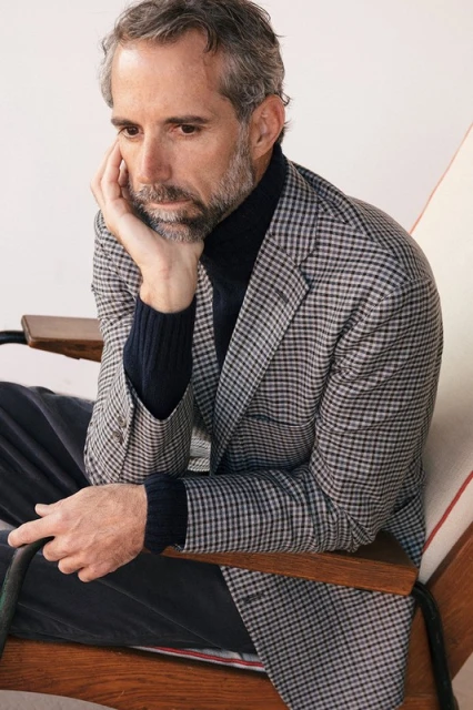 Portrait of a middle-aged man with salt and pepper hair, wearing a houndstooth blazer and a navy turtleneck, sitting in a chair indoors.