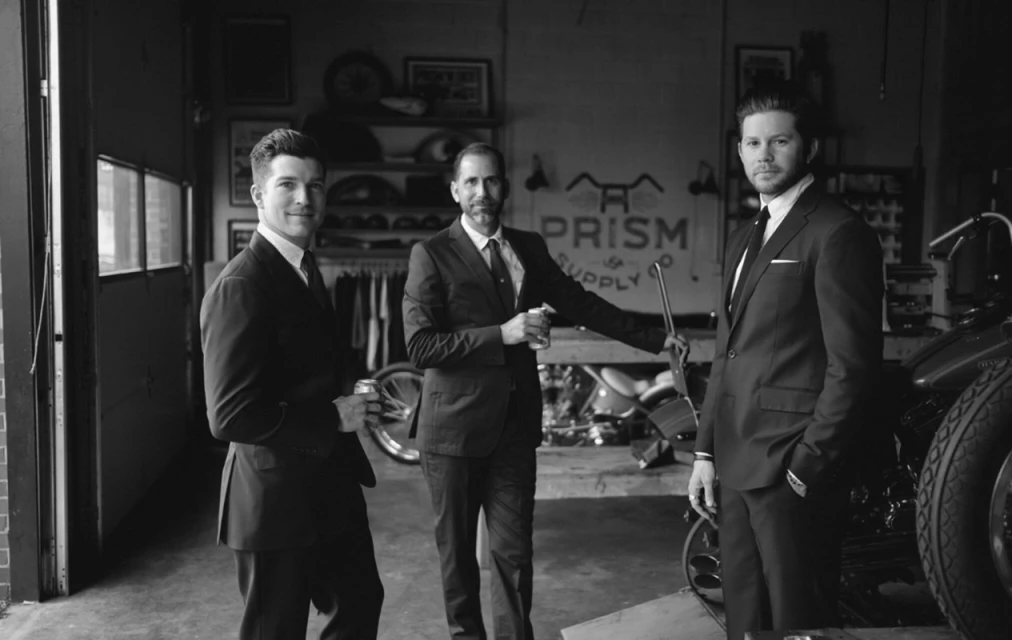 Three adult males pose indoors wearing suits, holding beverages next to vintage motorcycles in a black and white lifestyle photography. 