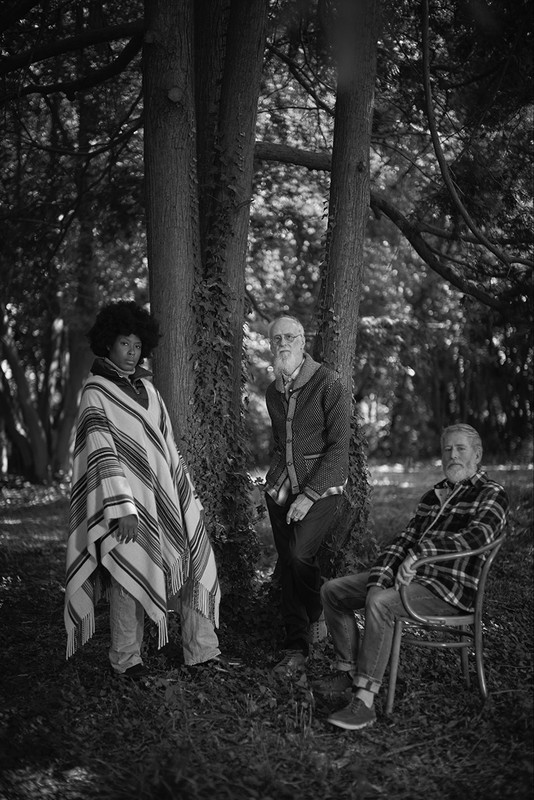 Three men pose outdoors in a black and white photograph. One stands wearing a poncho, one leans against a tree, and one sits on a chair.