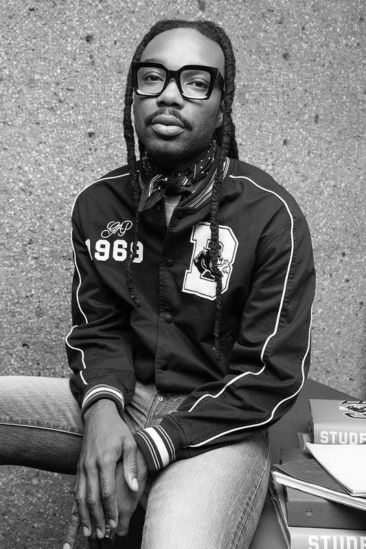 Black man wearing thick black frame glasses, a letterman jacket, and jeans seated outdoors against a textured concrete wall.