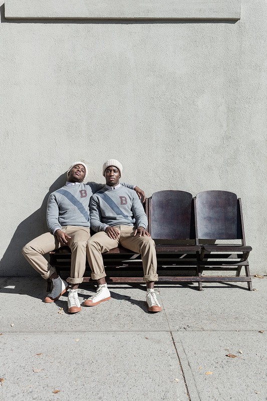 Two adult males in matching vintage sweaters and hats sit on a bench outside against a concrete wall on a sunny day.