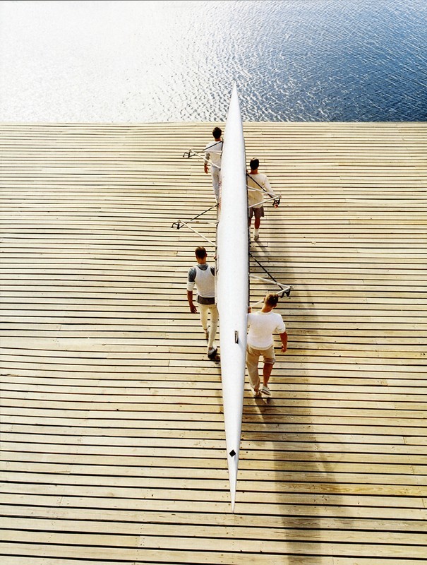 Overhead shot of four people carrying a white rowing scull along a wooden dock with a view of calm blue water in the background.