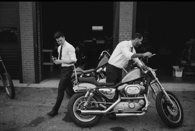 Two men in white shirts and dark trousers posing near motorcycles outside a garage
