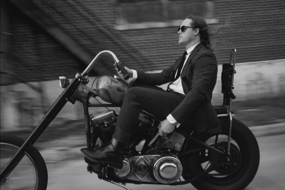 Monochrome shot of a man in a suit riding a chopper-style motorcycle on an urban street, showcasing a blend of formal attire and vintage style.