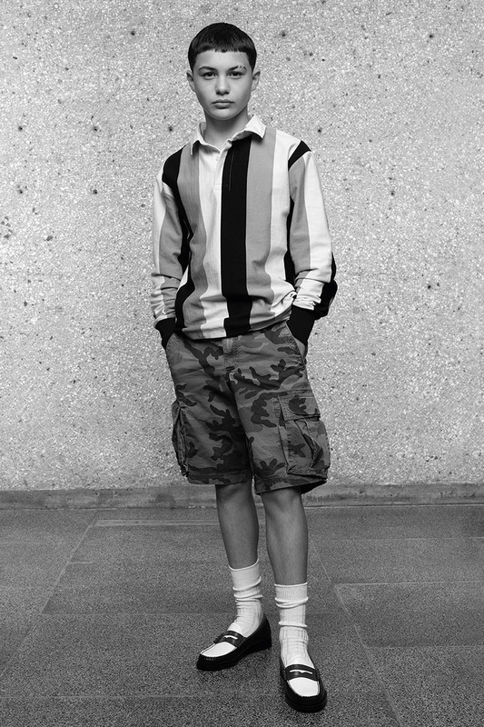 Teenager stands in front of a mottled concrete wall, wearing a striped rugby shirt and camouflage shorts, with white socks and loafers.