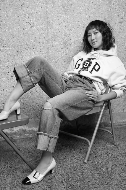 Black and white shot of a young woman in jeans and a hoodie sitting on a metal chair in front of a textured wall.