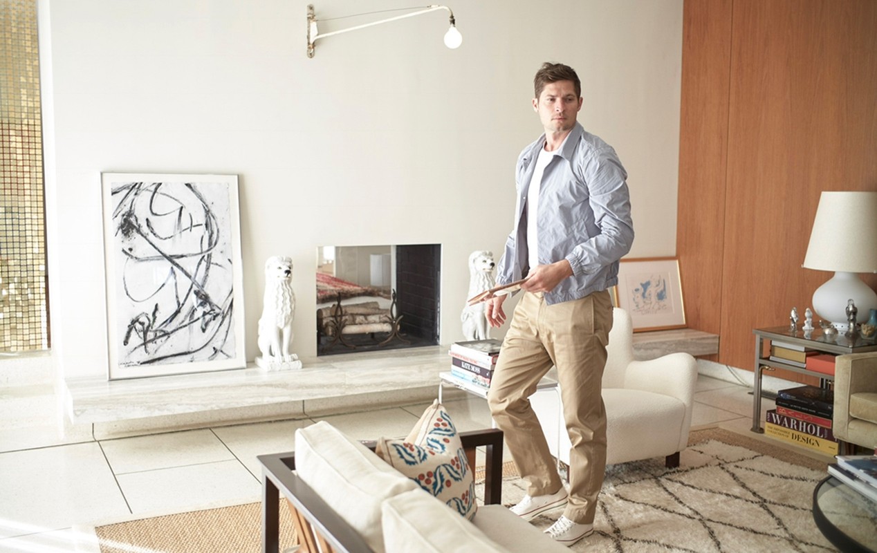 An adult male stands in a modern living room next to a fireplace and armchair, holding a thin wooden object in his hand.