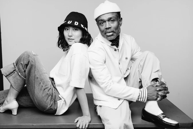 Monochrome studio portrait of an Asian woman and a Black man sitting back-to-back on a table styled by Scott Newkirk.