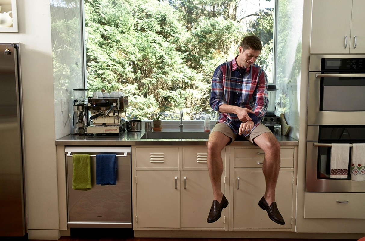 A blond man sits on a kitchen counter near a sink, wearing a plaid shirt and loafers, with a window and greenery in the background.