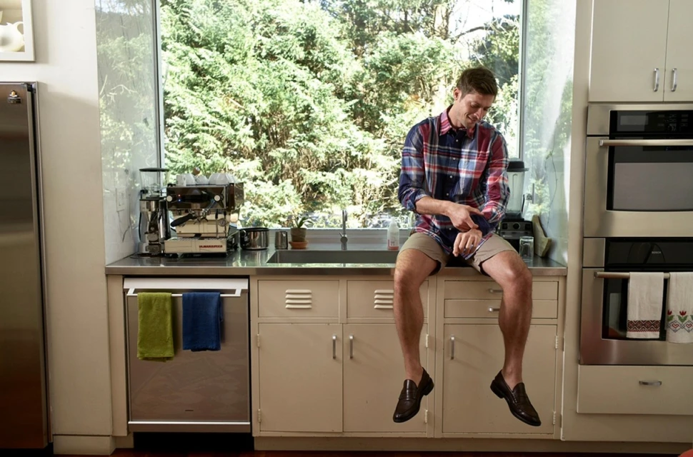 A blond man sits on a kitchen counter near a sink, wearing a plaid shirt and loafers, with a window and greenery in the background.