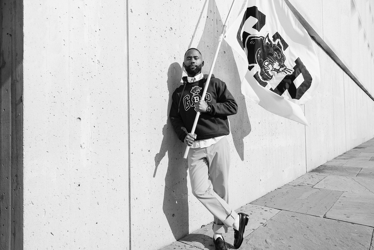 Black and white shot of a man holding a flag with a panther design, leaning against a concrete wall in an outdoor urban setting.