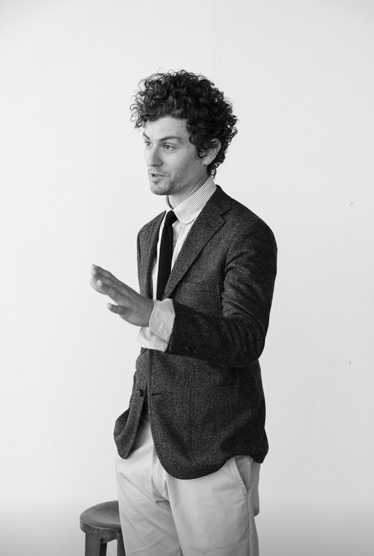 Monochrome portrait of a man with curly hair wearing a tweed blazer and tie against a white background