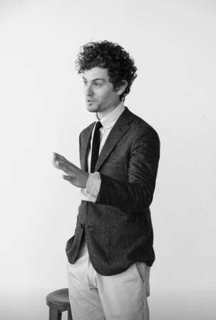 Monochrome portrait of a man with curly hair wearing a tweed blazer and tie against a white background