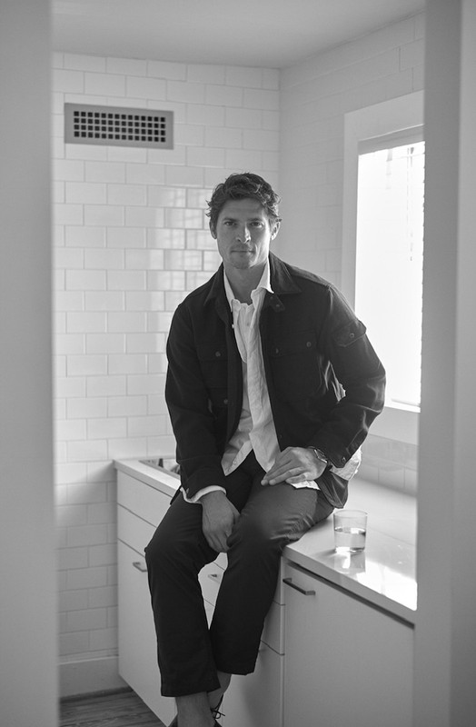 A fair-skinned adult male sitting on a white counter with a window and white tiled wall behind him, wearing a jacket and button-down shirt.
