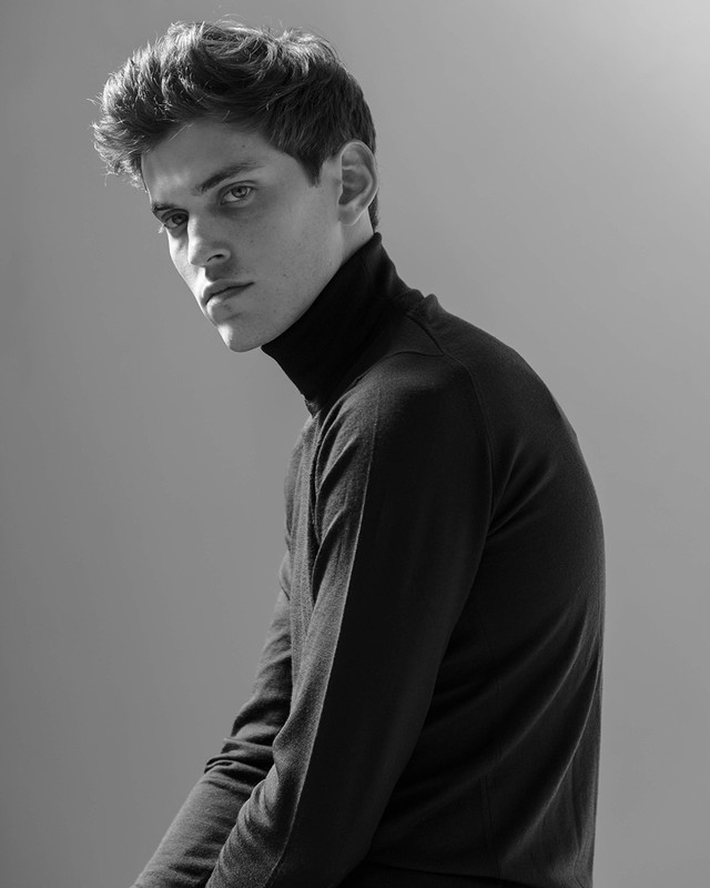 Monochrome portrait of a young man wearing a black turtleneck sweater looking over his shoulder against a grey background.