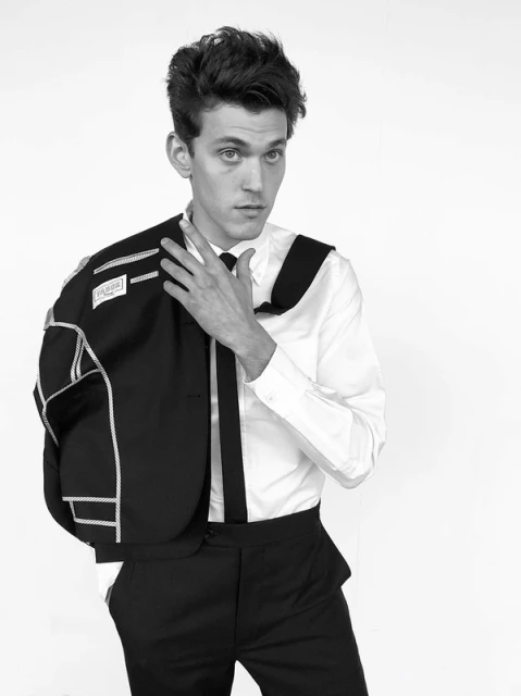 Black and white portrait of a young adult male wearing a white shirt and black dress pants, holding a suit jacket over his shoulder.