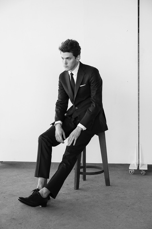 A man in a black suit and dark tie sits on a wooden stool against a plain studio backdrop.