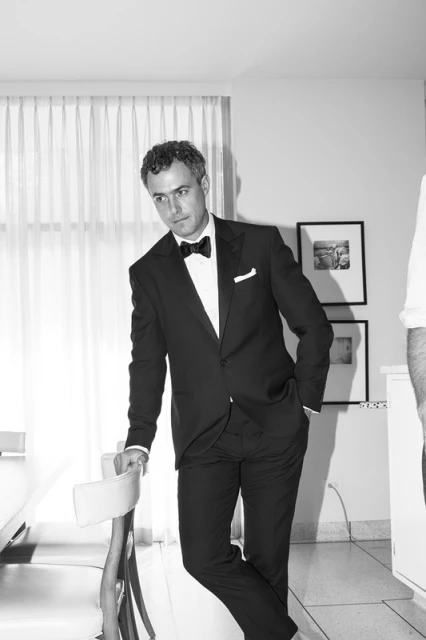 Black and white portrait of a man in a suit and bow tie leaning against a chair in an indoor setting with natural light.