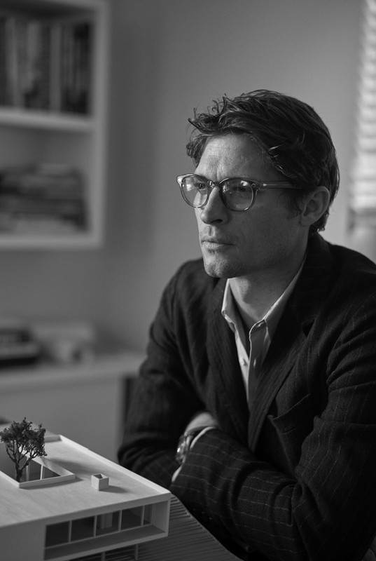 Black and white portrait of a man with glasses looking at a miniature building model on a table, with bookshelves in the background.
