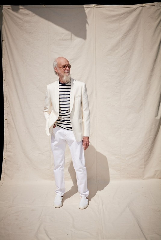Stylish elderly man in an all-white suit and striped shirt poses in front of an off-white backdrop, showcasing fashion and elegance.