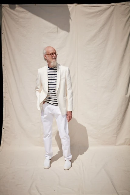 Stylish elderly man in an all-white suit and striped shirt poses in front of an off-white backdrop, showcasing fashion and elegance.