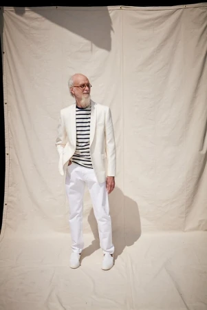 Stylish elderly man in an all-white suit and striped shirt poses in front of an off-white backdrop, showcasing fashion and elegance.