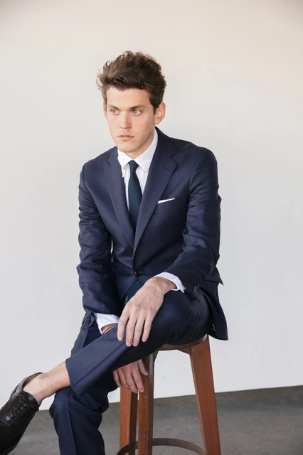A young, adult man sits on a wooden stool, wearing a navy suit, a white shirt, and black leather shoes, in a studio setting.