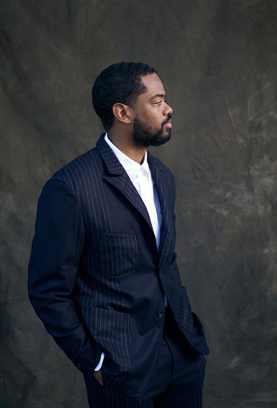 Portrait of a handsome adult Black man in a pinstripe suit, posing in a photo studio with a neutral expression and side view.