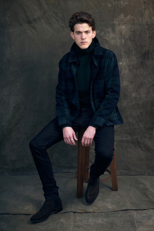 A young adult male in a dark green turtleneck and plaid jacket poses while sitting on a stool in a dimly lit, neutral studio.