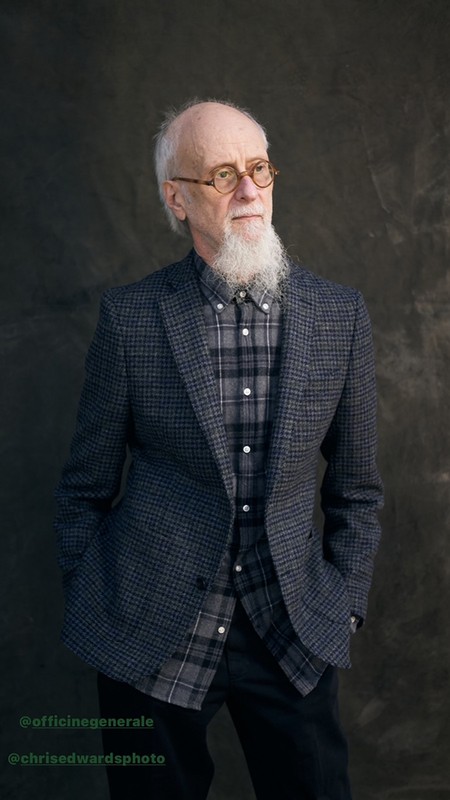 Elderly man with a white beard wearing round glasses, a plaid shirt, and a blazer against a dark textured background