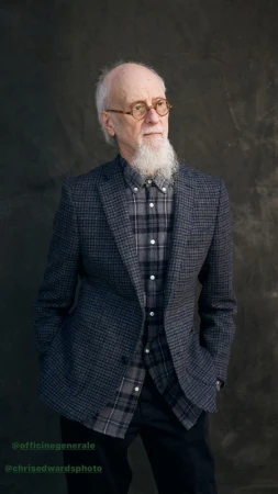 Elderly man with a white beard wearing round glasses, a plaid shirt, and a blazer against a dark textured background