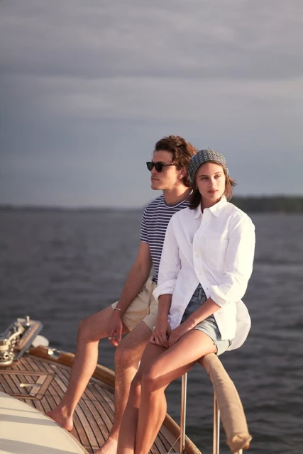 Couple sits on a wood boat, wearing casual clothing with a sea and cloudy sky in the background, creating a serene nautical scene.