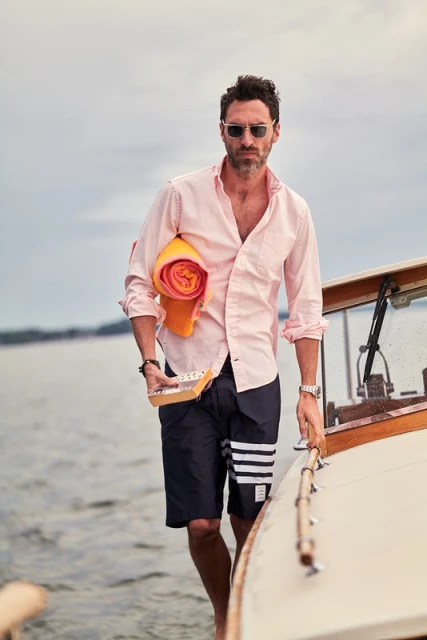 A man wearing sunglasses and a pink shirt stands on a boat, holding a yellow towel, on a cloudy day near the water.