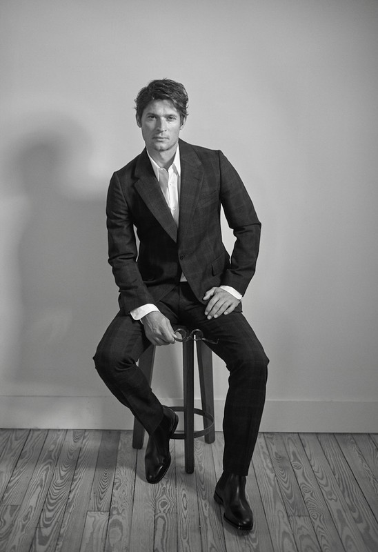 Black-and-white portrait of a man in a dark suit seated on a stool indoors with a light gray background and light-toned wood floor.