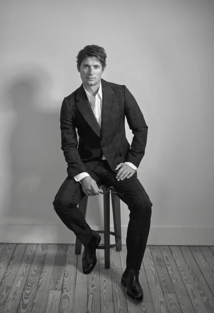 Black-and-white portrait of a man in a dark suit seated on a stool indoors with a light gray background and light-toned wood floor.