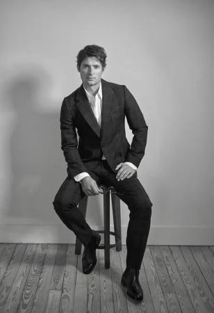 Black-and-white portrait of a man in a dark suit seated on a stool indoors with a light gray background and light-toned wood floor.