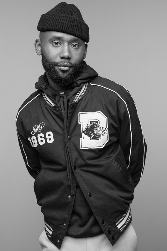Black and white portrait of a stylish Black male wearing a black knit cap and a letterman-style jacket with an embroidered design.