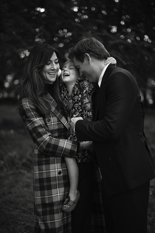 Black and white portrait of a smiling family of three hugging outdoors in a park with trees in the background.